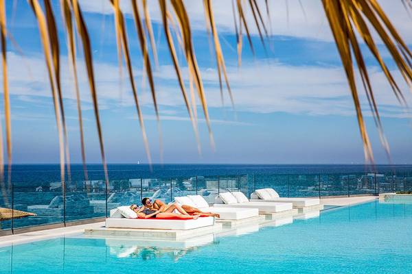 Zwei Personen liegen auf weißen Liegen mit roten Auflagen im Hotelpool mit Blick auf den Ozean.