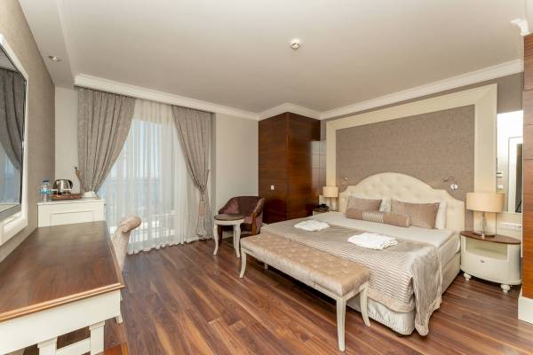 Innenansicht eines eleganten Hotelzimmers im Sunis Efes Royal Palace Resort & Spa mit Doppelbett, Nachttischen, Spiegel und Schreibtisch.