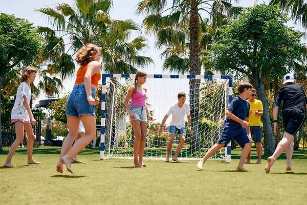 Mehrere Kinder spielen Fussball auf einem Kunstrasenplatz im Freien, vor einem Tor.