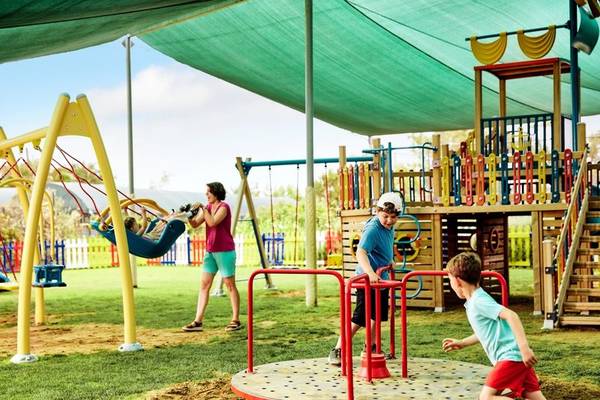 Familie auf dem Spielplatz im Magic Life Jacaranda Hotel, Kinder spielen auf Schaukeln und Karussell.