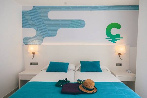 Zimmer im Calimera Balansat Resort mit zwei Betten, blauer Bettwäsche, einem Hut und Handtaschen auf dem Bett.