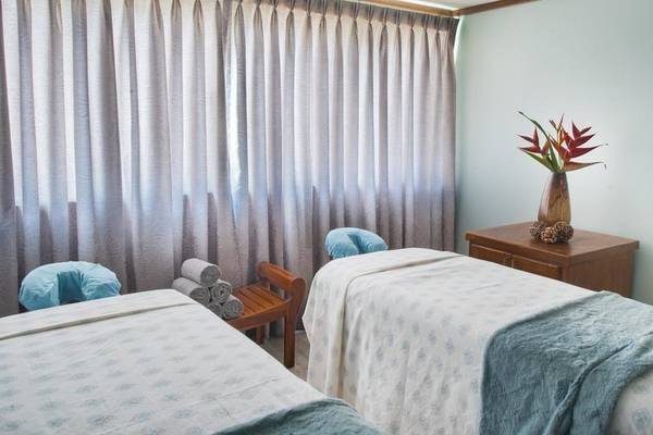 Massage-Behandlungsraum im Sands Barbados mit zwei Massageliegen, grauen Vorhängen und einem Tisch mit Blumenvase.