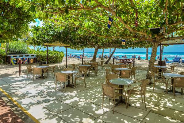 Außenterrasse des Coconut Court Beach Hotel mit Tischen und Stühlen unter Bäumen, Blick auf den Strand und das Meer.