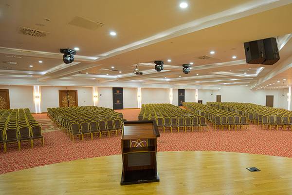 Saal im Alan Xafira Deluxe Resort & Spa mit vielen Stühlen, einem Podium und Lautsprechern an der Decke.