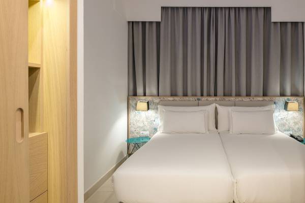 Doppelbett-Zimmer im Plus Fariones Suite Hotel mit weißen Laken und Kissen, grauen Vorhängen und blauen Nachttischen.