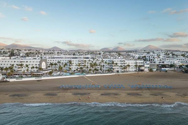 Außenansicht des Plus Fariones Suite Hotel mit Strand, blauen Liegestühlen und weißen Gebäuden.