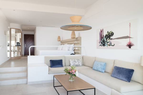 Innenraum des Plus Fariones Suite Hotels mit Wohnzimmersofa, Tisch, Blumenvase und Schlafzimmerbereich.