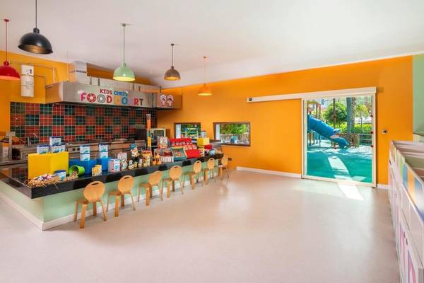 Kinderkoch-Essensbereich im Kirman Leodikya Resort mit gelben Wänden, Holzstühlen und Spielplatz im Hintergrund.