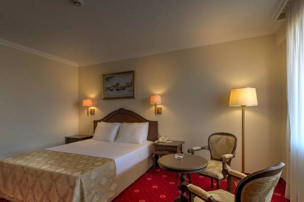 Ein elegantes Hotelzimmer im Ducale Lara mit einem Doppelbett, zwei Sesseln, einem kleinen Tisch und einer Wand mit zwei Lampen und einem Gemälde.