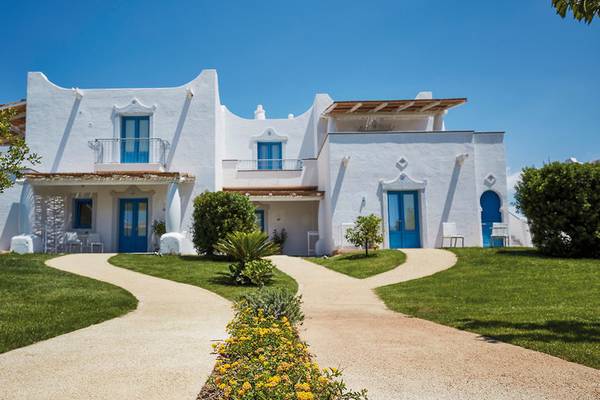 Außenansicht des weißen VOI Marsa Siclà Essentia Hotels mit blauen Türen und grünem Rasen.