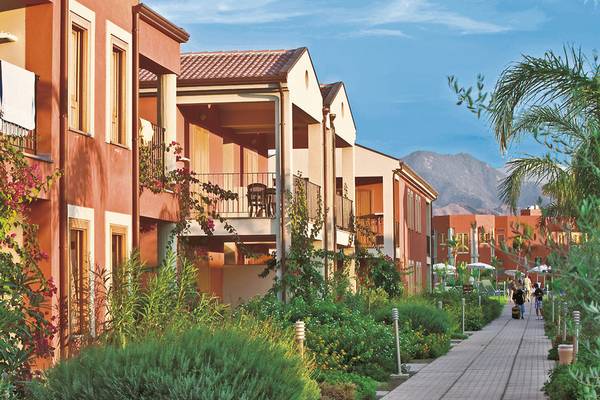 Außenansicht des VOI Baia di Tindari Resorts mit orangefarbenen Gebäuden und Balkonen, umgeben von Pflanzen und einem gepflasterten Weg.