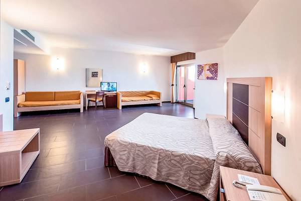 Innenansicht eines Hotelzimmers im VOI Baia di Tindari Resort mit Doppelbett, Sofas, Schreibtisch, Fernseher und Balkon.