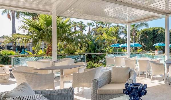 Terrasse im Acacia Palace Hotel mit weißen Möbeln, blauen Sonnenschirmen und Blick auf einen Pool und Palmen.