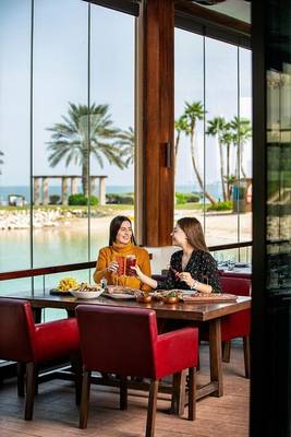 Zwei Frauen im Sheraton Doha Resort Convention Hotel, sitzend am Tisch mit Essen und Getränken vor einer Panorama-Seeansicht.