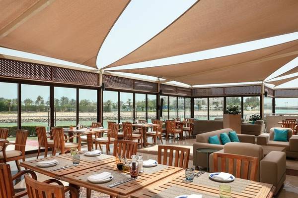 Innenbereich des Restaurants im Grand Hyatt Doha Hotel & Villas mit Holztischen und Stühlen, Sonnenschirmen und Blick auf den Ozean.