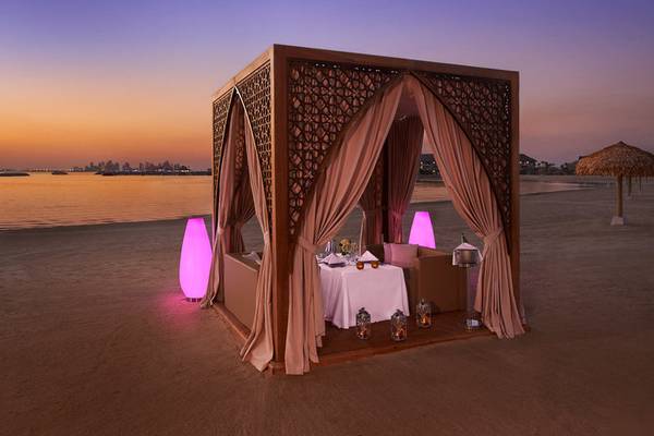 Strand-Gazebo mit Tisch und Sesseln, umgeben von pinken Leuchten und Vorhängen, bei Sonnenuntergang.