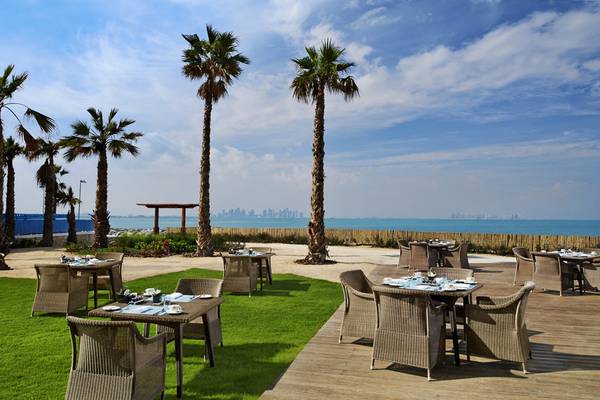 Außenbereich des Banana Island Resort Doha by Anantara mit Tischen, Stühlen und Palmen, Blick aufs Meer.