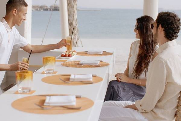 Drei Gäste sitzen am Tisch am Strand des Banana Island Resorts Doha mit Getränken und Blick aufs Meer.