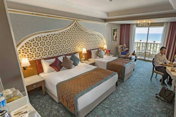 Innenansicht eines Zimmers im Royal Taj Mahal Hotel mit zwei Betten, einem Mann am Schreibtisch und einer Frau auf dem Balkon.