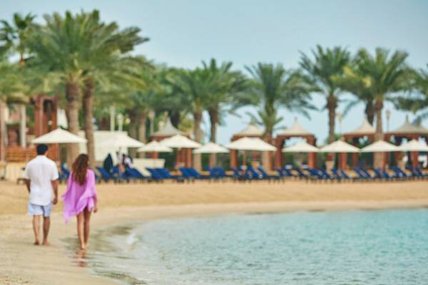 Ein Paar geht am Strand des Four Seasons Hotel Doha entlang, umgeben von Palmen und Sonnenschirmen.