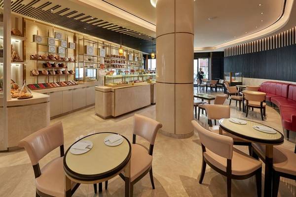 Innenbereich des Mandarin Oriental Doha Hotels mit modernen Möbeln, Esstischen und Stühlen, einer Theke mit Backwaren und einer Person in der Nähe.