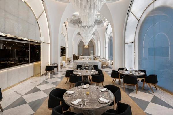 Innenansicht des Raffles Doha Restaurants mit Marmorboden, schwarzen Stühlen und runden Tischen, einem großen Kronleuchter und einer Küche im Hintergrund.
