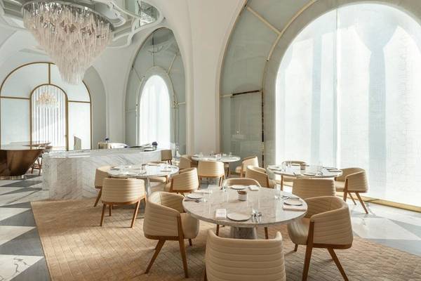 Innenansicht des Raffles Doha Restaurants mit runden Marmortischen, goldenen Kronleuchtern und großen bogenförmigen Fenstern.