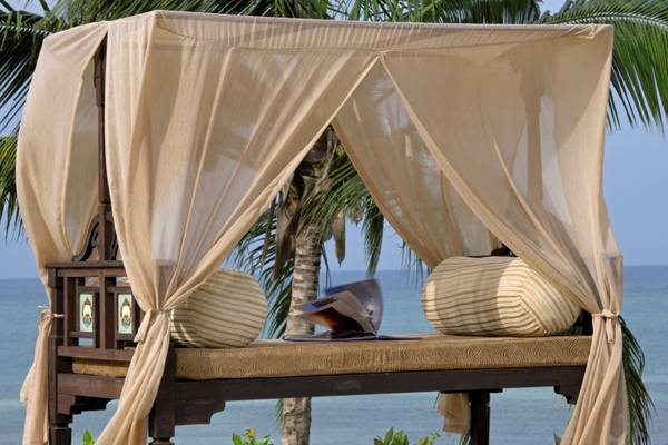 Doppelbett mit Vorhang am Strand mit Blick auf das Meer und Palmen im Hintergrund.