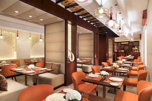 Innenansicht des Restaurants im Souq Al Wakra Hotel Qatar By Tivoli mit orangefarbenen Stühlen und Tischen, weißen Tellern und Blumenarrangements.