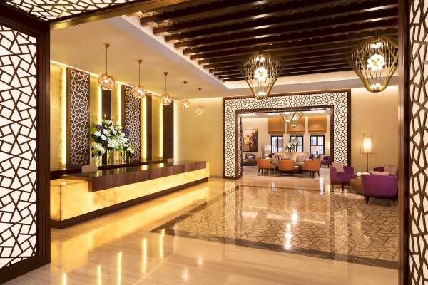 Lobby des Souq Al Wakra Hotel Qatar By Tivoli mit goldenen Wänden und leuchtenden Kronleuchtern.