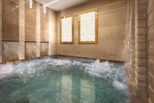 Schwimmbecken im Souq Al Wakra Hotel Qatar By Tivoli mit Wasserstrahlen und goldenen Fenstern.