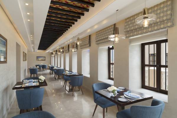 Innenansicht des Restaurants im Souq Al Wakra Hotel Qatar By Tivoli mit blauen Stühlen und Tischen, Deckenlampen und Fenstern.