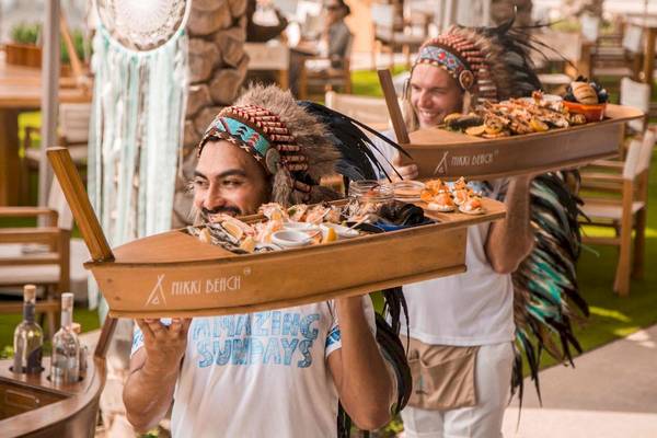 Mitarbeiter des Nikki Beach Resort & Spa Dubai tragen Speisen auf einem Holztablett.