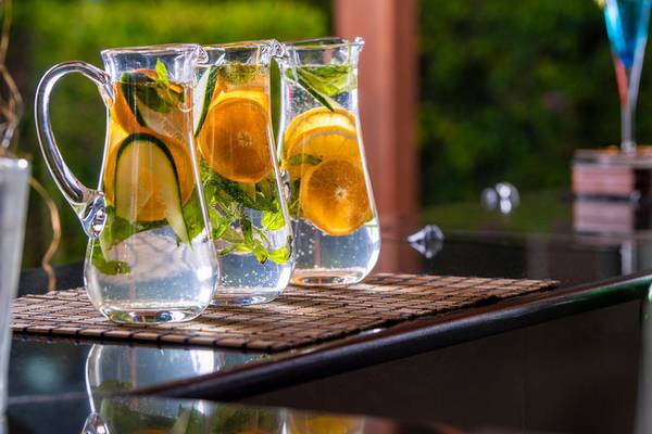 Drei Glaskruecke mit Wasser, Orangenscheiben, Gurkenscheiben und Minze auf einem Tisch.