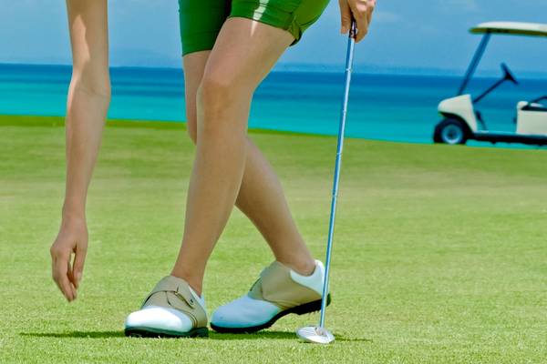 Golfspieler in grünen Shorts am Strand, ein Golfschläger haltend, mit einem Golfwagen im Hintergrund.