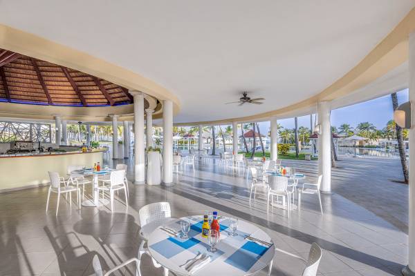 Innenbereich des Paradisus Varadero Resort & Spa mit mehreren weiß gedeckten Tischen und Stühlen, einem Blick auf einen Swimmingpool und tropischen Bäumen im Hintergrund.