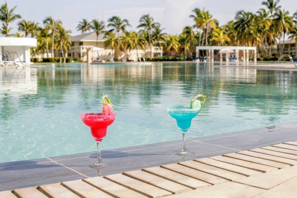 Zwei Cocktails mit rotem und blauem Getränk stehen am Rand eines Pools mit Palmen im Hintergrund.