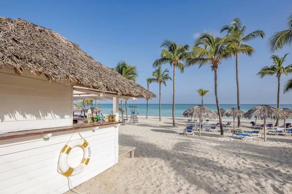 Strandszene mit einem kleinen weißen Gebäude, einem Strohdach und Palmen im Hintergrund.