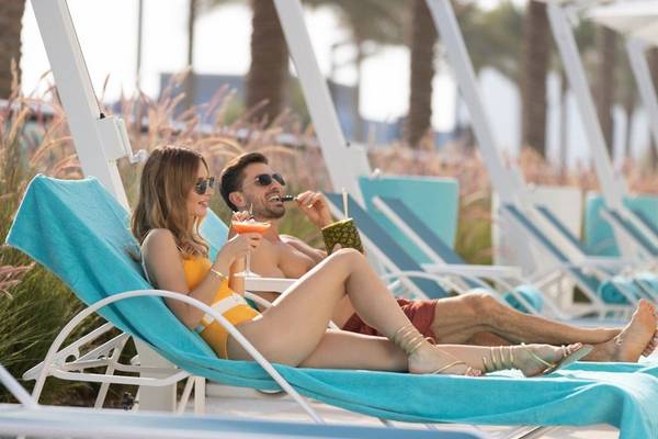 Paar im Hilton Dubai Palm Jumeirah auf Liegen am Pool, mit Cocktails, Sonnenbrillen und lächelnd.