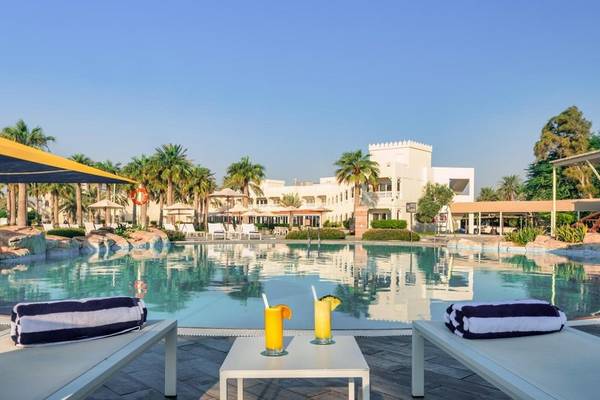 Außenpoolbereich des Sealine Beach, A Murwab Resort mit zwei Liegen, Getränken und einer Palmenlandschaft im Hintergrund.
