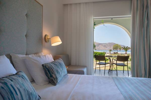 Ein Hotelzimmer mit einem Bett, Kissen, Lampen und einem offenen Balkon mit Blick auf den Strand.