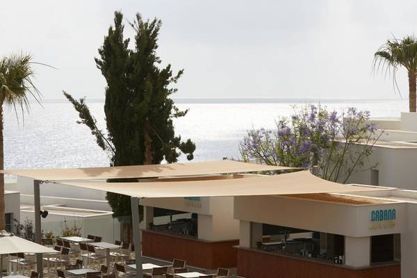 Außenbereich des Mare Paphos Hotels mit Blick auf den Ozean, Palmen und einer Terrasse mit Sonnenschirmen und Tischen.