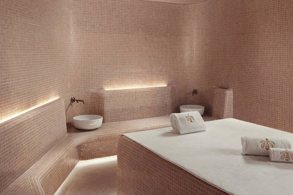 Spa-Behandlungsraum im St. Regis Al Mouj Muscat Resort mit gefliesten Wänden, Sitzbänken, weißen Waschbecken und Handtüchern.