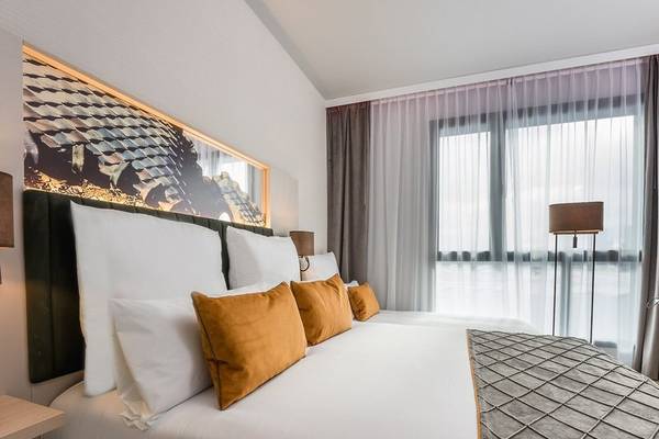 Schlafzimmer im Leonardo Royal Hotel Barcelona Fira mit zwei Betten, weißen und orangefarbenen Kissen, Kopfteil und großer Fensterfront.