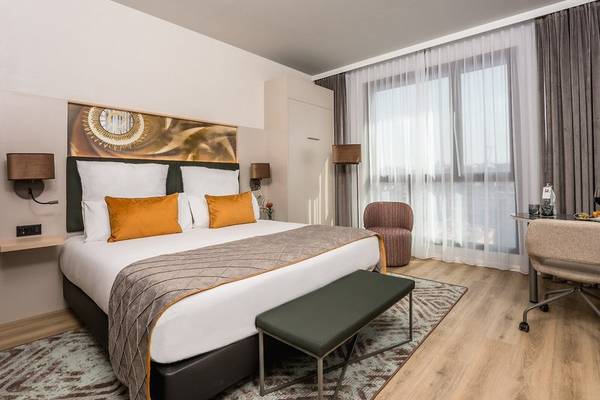 Schlafzimmer im Leonardo Royal Hotel Barcelona Fira mit großem Bett, weißen und orangefarbenen Kissen, gemusterter Decke, Nachttischen und Lampen.