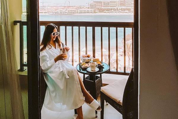 Frau im Bademantel auf Balkon im Traders Hotel Qaryat Al Beri mit Blick auf das Meer.