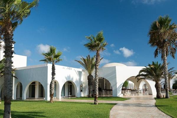 Weißes Hotelgebäude mit Bogenfenstern, umgeben von Palmen und Rasen, heller Himmel im Hintergrund
