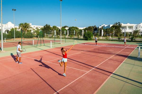 Tennisplatz im Hotel Iliade Djerba mit Spielern und Gebäuden im Hintergrund.