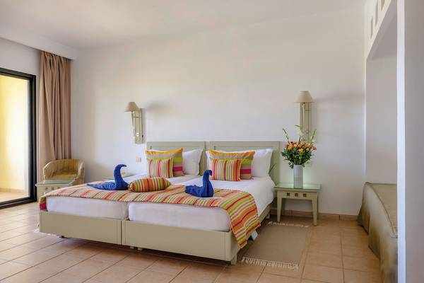 Hotelzimmer im Aldiana Club Djerba Atlantide mit zwei Betten, bunten Kissen, Tisch mit Blumenvase, und offenem Fenster.