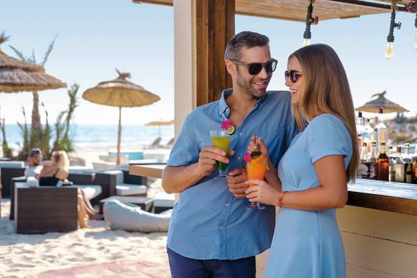 Ein Paar steht lächelnd an einer Strandbar und hält Cocktails in der Hand.